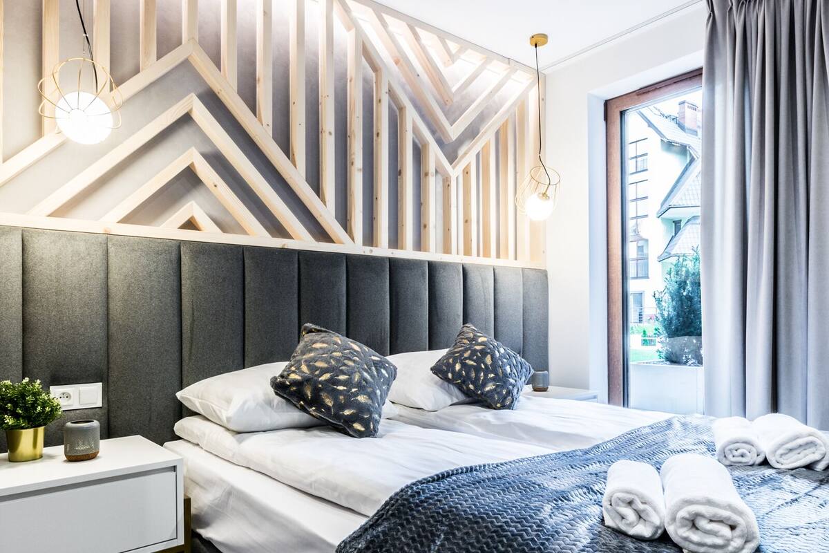 Una habitación de hotel moderna con una cama grande, cabecera con diseños geométricos, una mesita de noche con una planta y vistas al exterior a través de una ventana.
