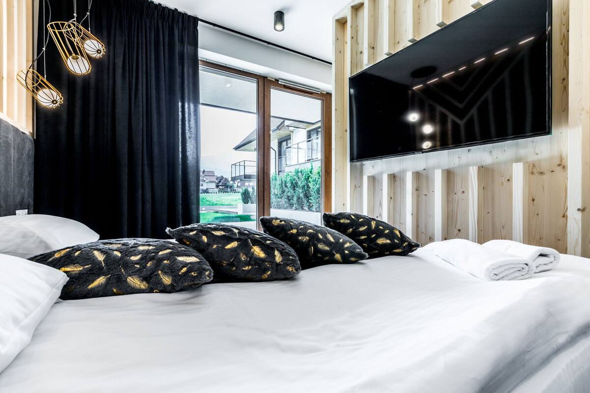 Habitación de hotel moderna con una cama grande, almohadas estampadas en negro y dorado, un televisor de pantalla plana montado en una pared de madera y vistas al exterior a través de amplias ventanas.