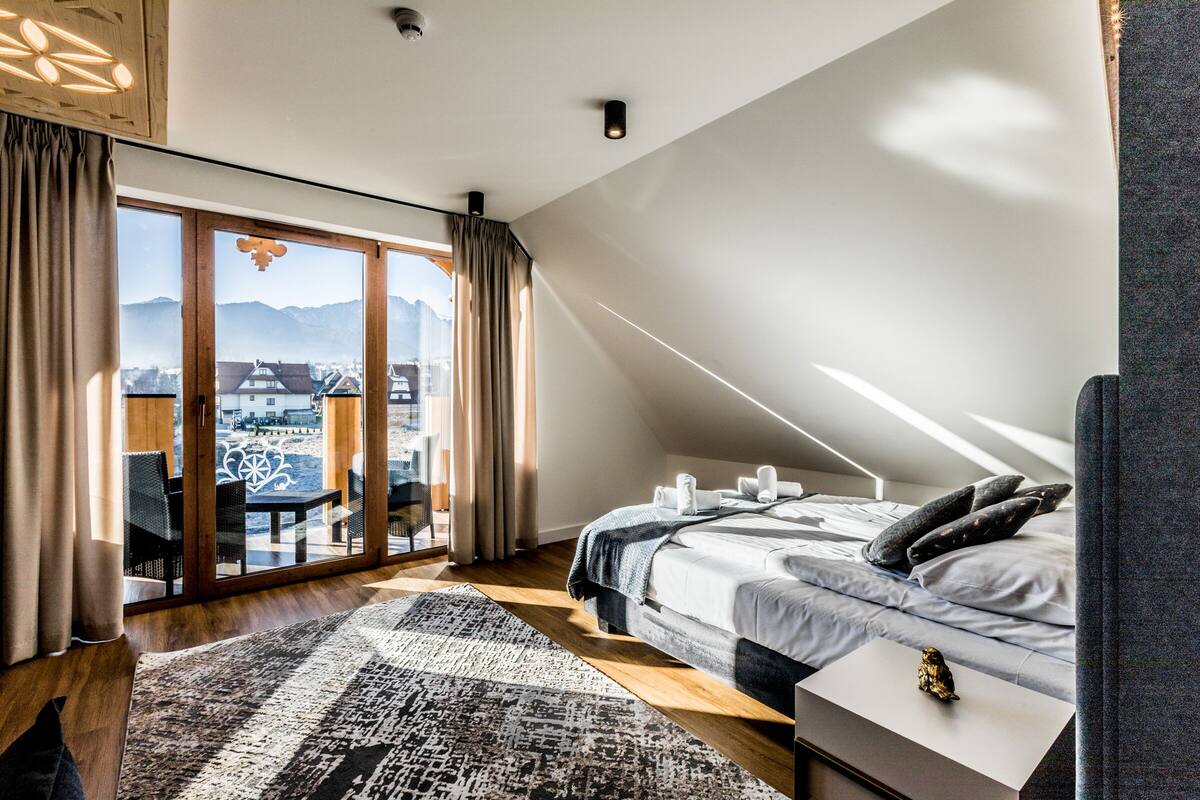 Un dormitorio con una cama grande, vistas de la ciudad a través de ventanales del suelo al techo y un balcón con mobiliario exterior.