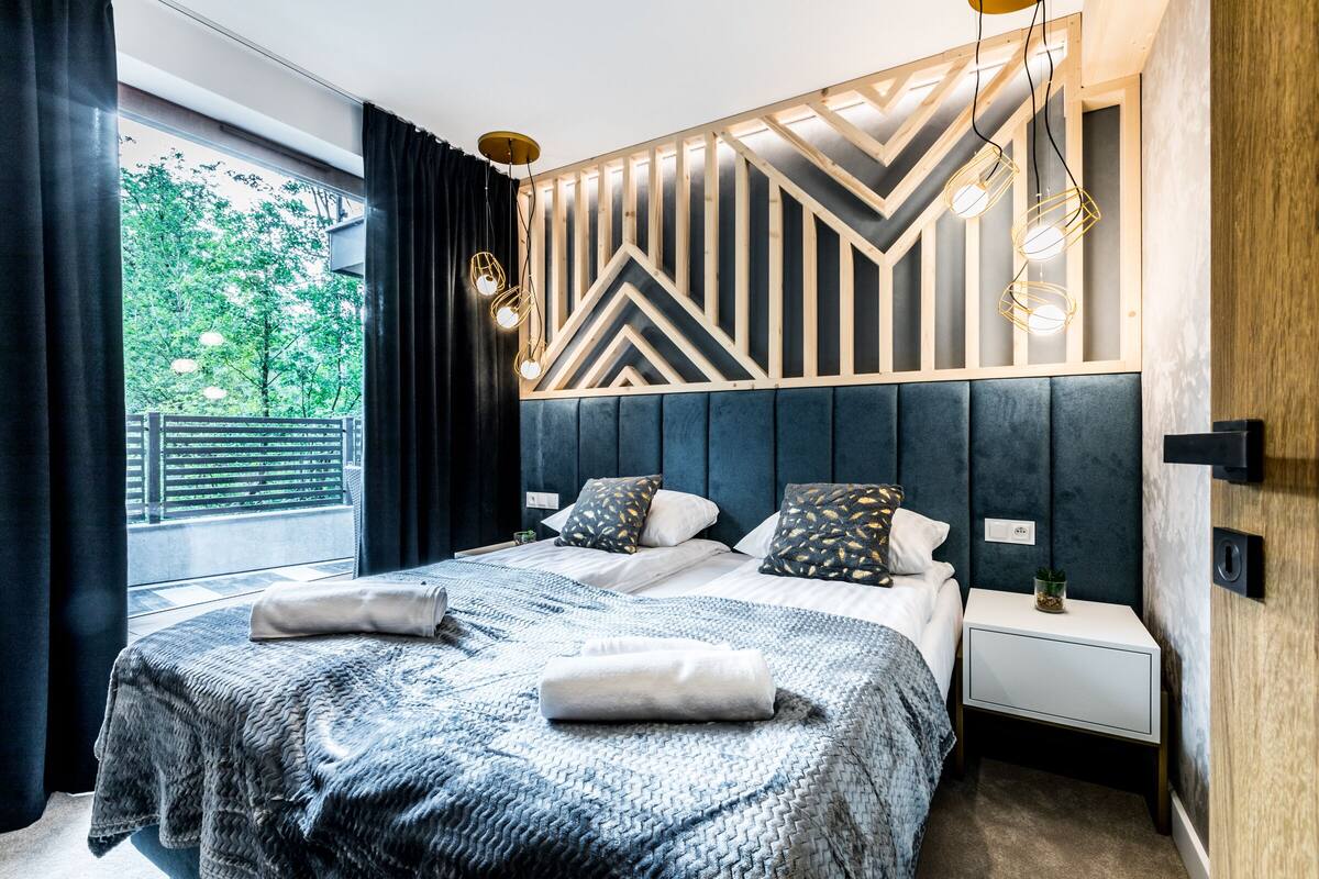 Un dormitorio moderno con una cama grande, cabecero con diseños geométricos, mesitas de noche y vistas al exterior con vegetación.