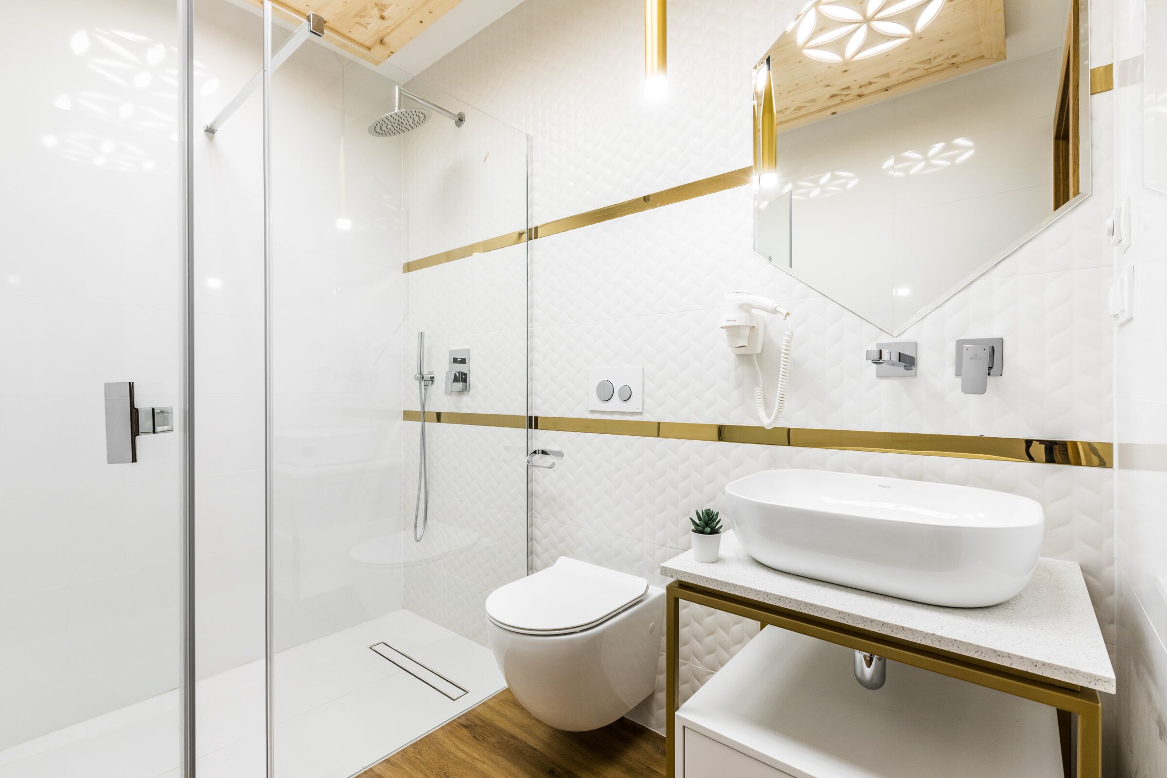 Een moderne badkamer met een douchecabine van glas, een toilet, een wastafel met gouden lijst en een spiegel met een geometrische lamp.