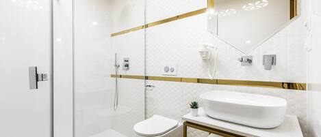 Een moderne badkamer met een douchecabine van glas, een toilet, een wastafel met gouden lijst en een spiegel met een geometrische lamp.