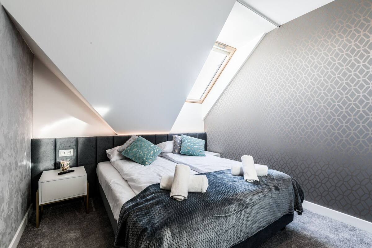 Un dormitorio moderno con techo inclinado, una cama con manta gris, dos almohadas y una mesita de noche con lámpara.