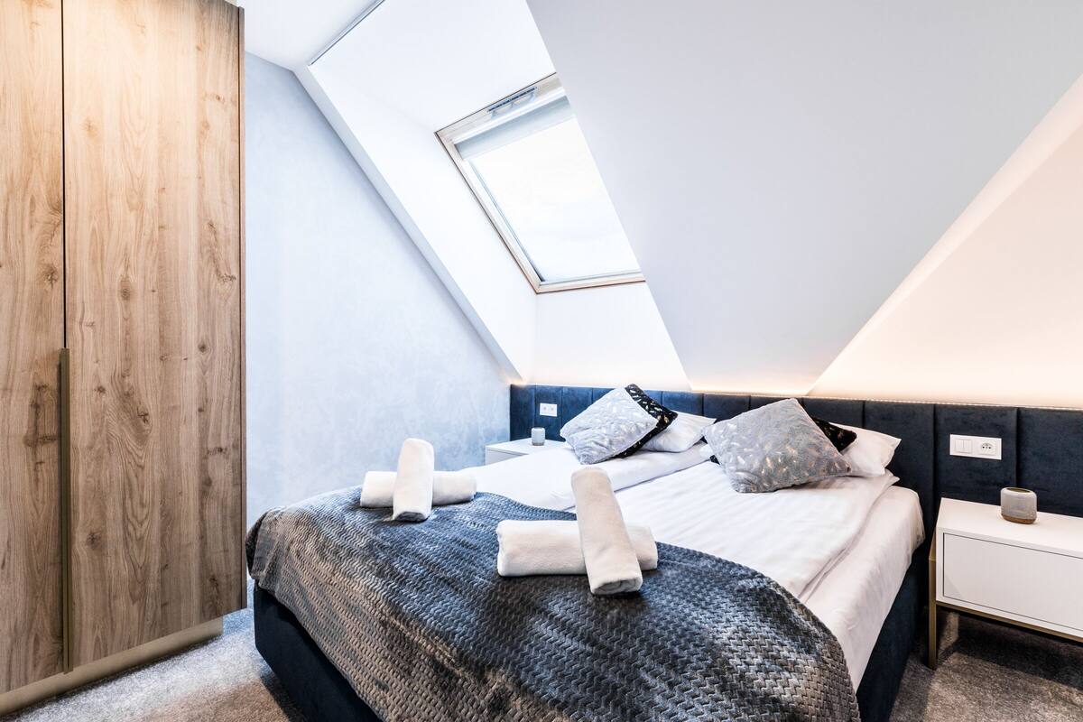 Un dormitorio moderno con techo inclinado, tragaluz, cama bien hecha con toallas, mesita de noche y mesilla.
