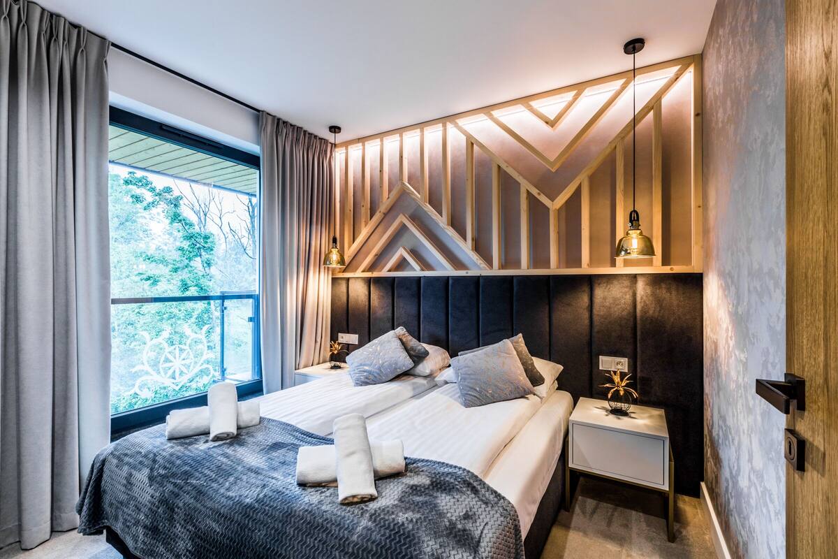 Un dormitorio moderno con una cama grande, cabecera con diseños geométricos, mesitas de noche con lámparas y vista a árboles por la ventana.