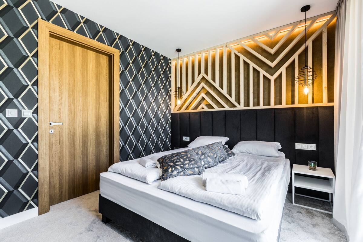 Un dormitorio moderno con pared de diseño geométrico, puerta de madera, cama con ropa blanca y cabecero empotrado con iluminación integrada.