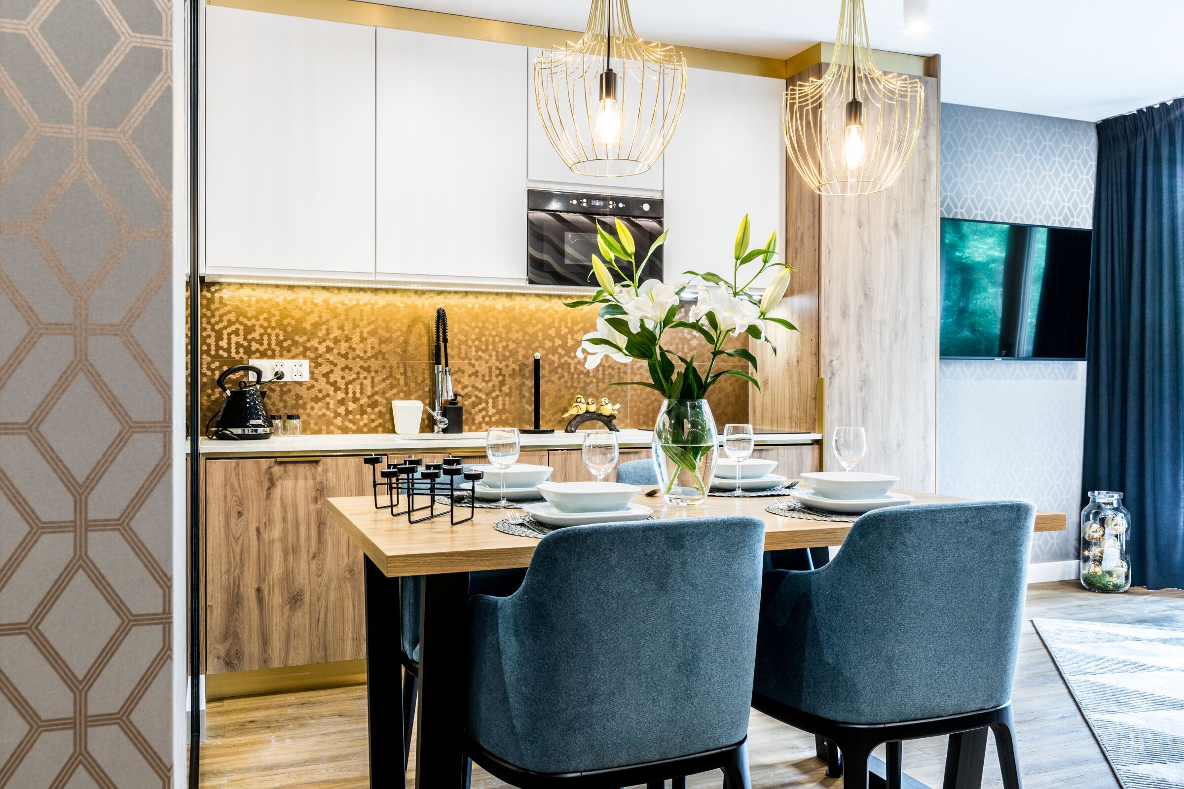 Ein moderner Essbereich mit einem Holztisch, gedeckt für zwei Personen, einer Vase mit Blumen und einer Küche im Hintergrund.