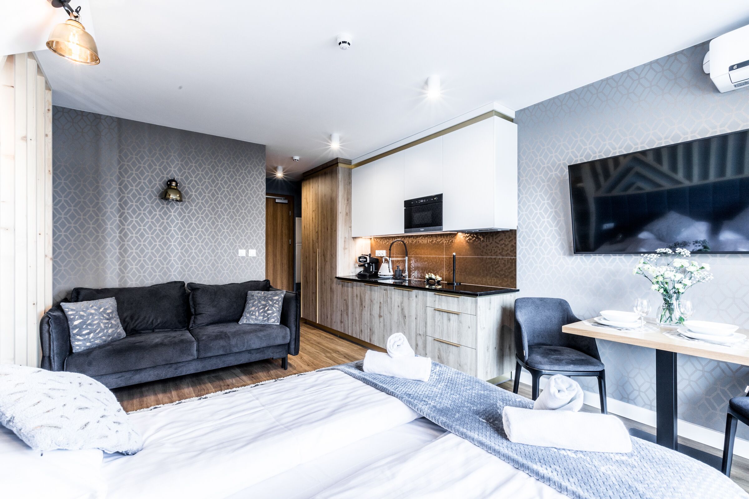 Quarto de hotel moderno com cama, sofá, mesa de jantar com cadeiras, cozinha e TV fixada na parede.