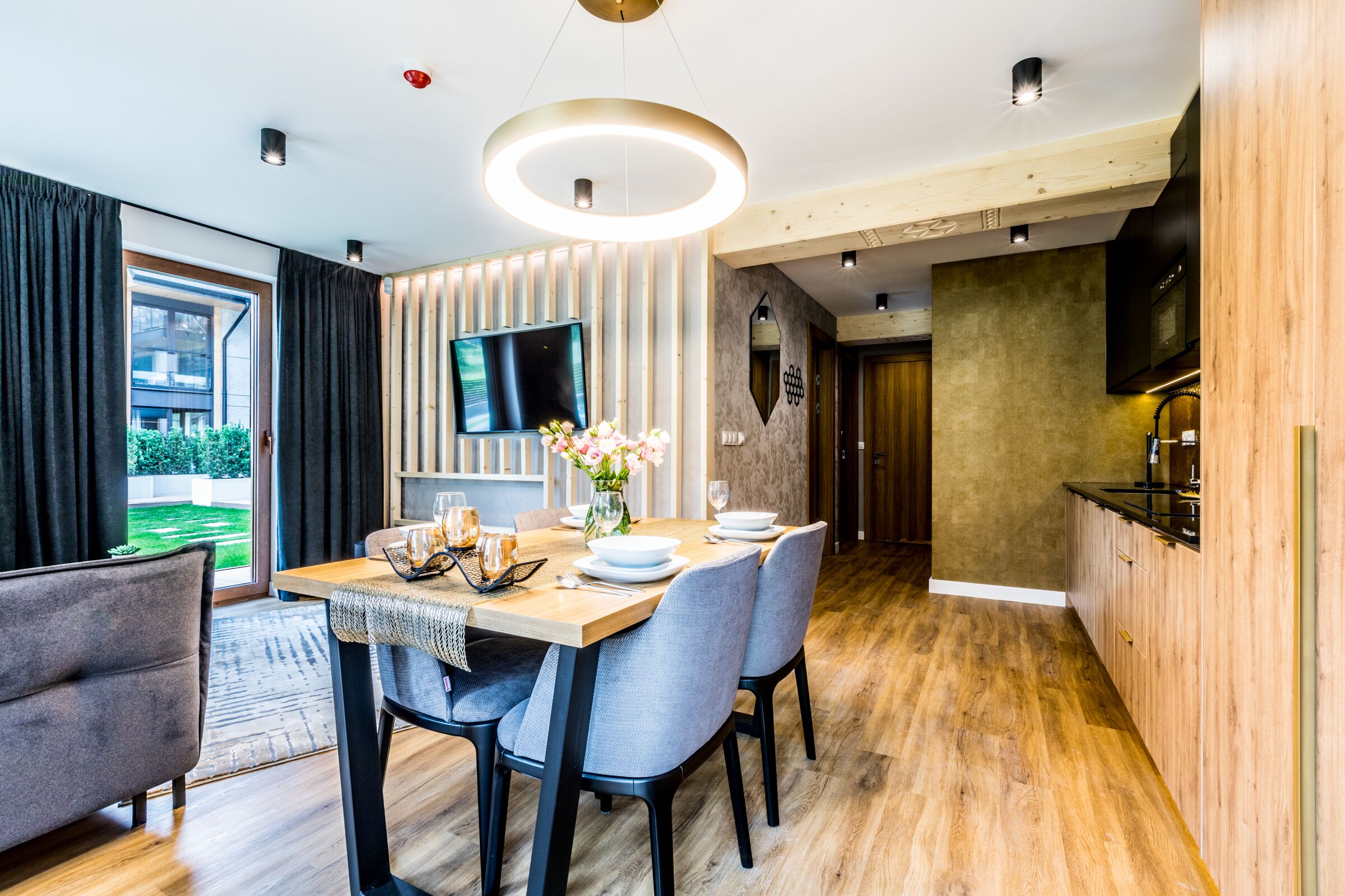 A modern dining area with a wooden table, grey upholstered chairs, and a flat-screen TV mounted on the wall.