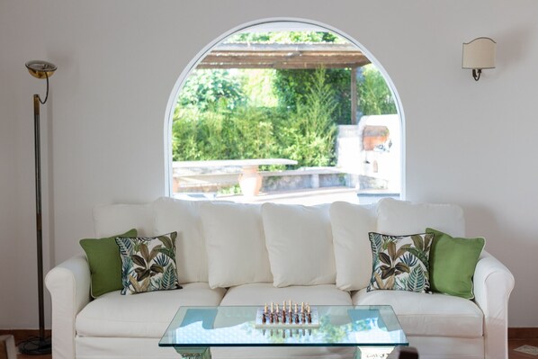 Un canapé blanc agrémenté de coussins à motifs verts et blancs, une table basse en verre et une vue sur la verdure à travers une grande fenêtre en arc de cercle.