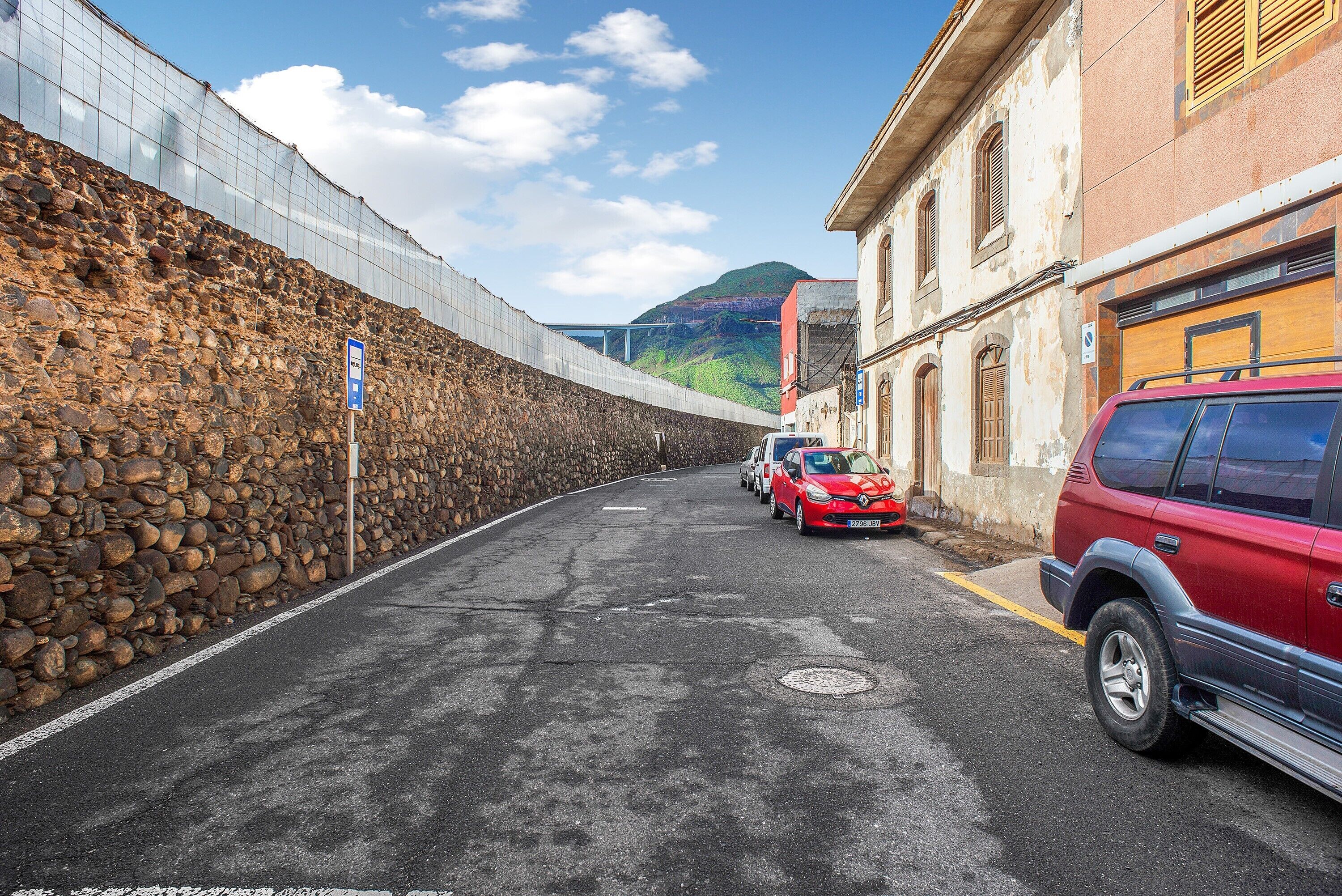 A street with a stone wall, parked cars, and a building with a red roof.