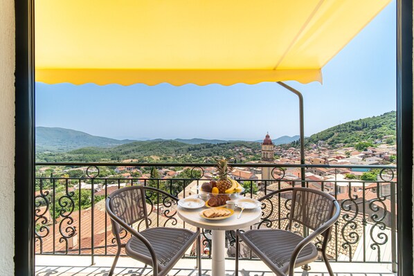 A balcony with a table set for two, overlooking a scenic town with hills and a church.