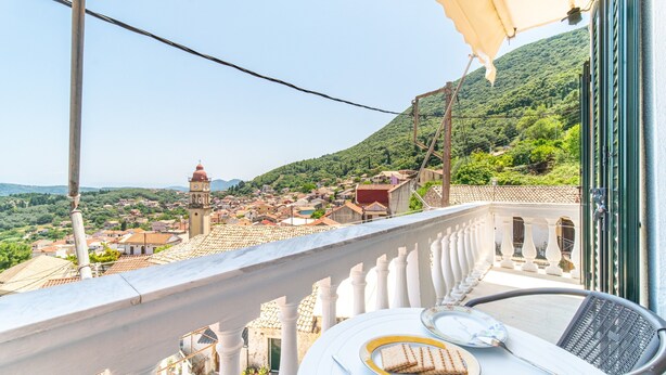 Un balcone con un tavolo apparecchiato per due, affacciato su una città con il campanile di una chiesa e dolci colline verdi.