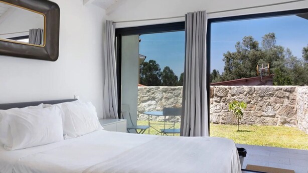 Une chambre avec un lit, une petite table et une chaise, une grande fenêtre avec des rideaux, et une vue sur un mur de pierre et la verdure à l’extérieur.