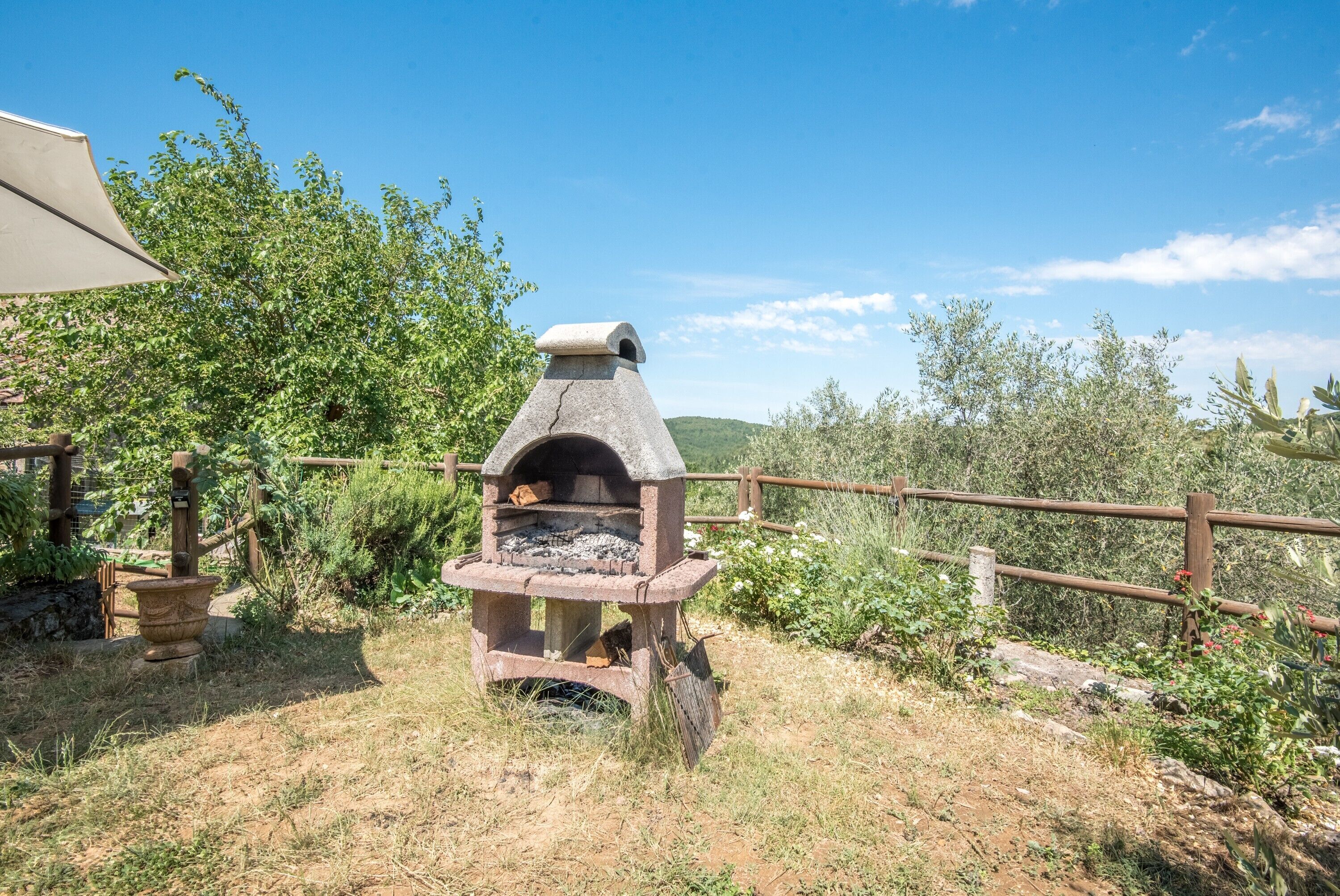 Forn de pedra a l'aire lliure amb xemeneia, envoltat d'una tanca de fusta i vegetació.