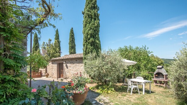 Un cadre extérieur rustique avec un bâtiment en pierre, une table en bois entourée de chaises, un four en briques et une variété de plantes, notamment de grands cyprès et des plantes à fleurs.