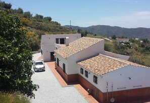 Un edificio de dos pisos con techo de tejas, un coche estacionado al frente y un paisaje montañoso al fondo.