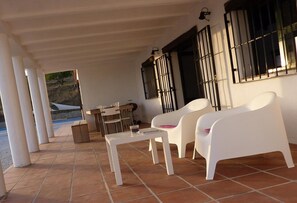 Un patio techado con sillas de plástico blanco, una mesita y una ventana con barrotes negros.
