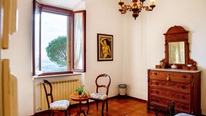 Een kamer met een houten dressoir, een stoel, een tafel met een plant, een radiator, een raam met gordijnen, een kroonluchter en een lijst met een afbeelding aan de muur.