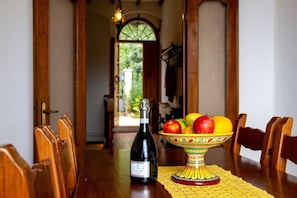 Una mesa de comedor con una botella de vino, un cuenco de fruta y sillas de madera.