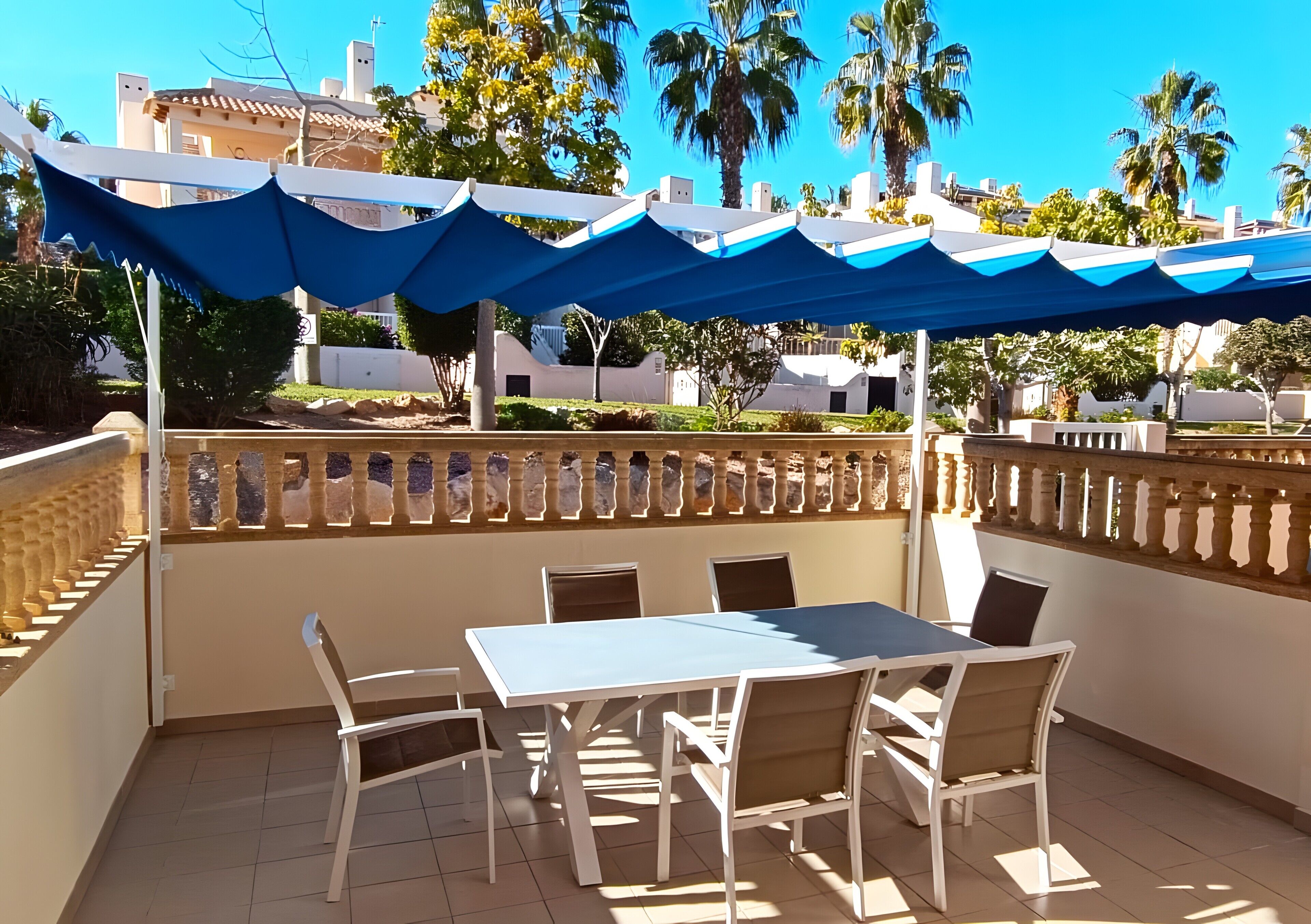 Een terras met een tafel en stoelen, een blauw zonnescherm en uitzicht op palmbomen en gebouwen.