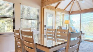 Un coin repas avec une table en verre et des chaises en bois, de grandes fenêtres équipées de stores et un ventilateur de plafond.