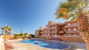 A swimming pool surrounded by a tiled deck, a building with balconies, and palm trees. - Holiday Home 'Vista Mar' with Sea Views, Shared Pool and Wi-Fi (Puertito de Güímar)