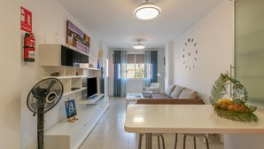 A modern living room with a flat-screen TV, a dining table, and a sofa. There is a fire extinguisher on the wall and a fan in the corner. - Holiday Home 'Vista Mar' with Sea Views, Shared Pool and Wi-Fi (Puertito de Güímar)
