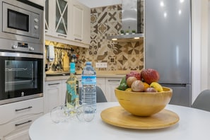 A kitchen with a microwave, oven, and refrigerator. There is a bowl of fruit on the table.