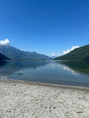 Un lago tranquilo con montañas al fondo, cielo despejado y una playa de guijarros.