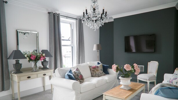 Ein Wohnzimmer mit weißem Sofa, einem Holztisch, einem weißen Sessel, einem Fernseher an der Wand und einer Kandelaber.