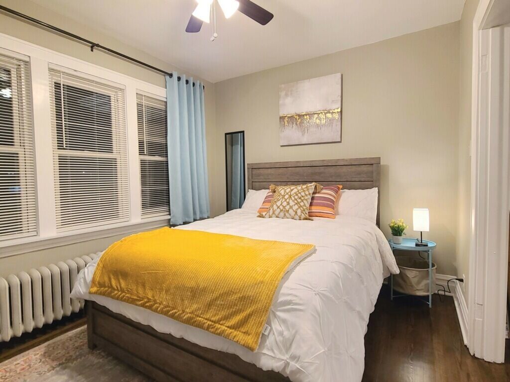 Une chambre à coucher avec un lit, une table de chevet, une lampe, un radiateur et une fenêtre avec des stores.