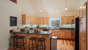 A modern kitchen with wooden cabinets, a central island with stools, a stainless steel refrigerator, and a chalkboard sign on the wall.