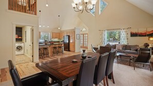 A spacious living area with a wooden dining table, black chairs, a sofa, and a coffee table. A kitchen with wooden cabinets and a refrigerator is visible in the background.