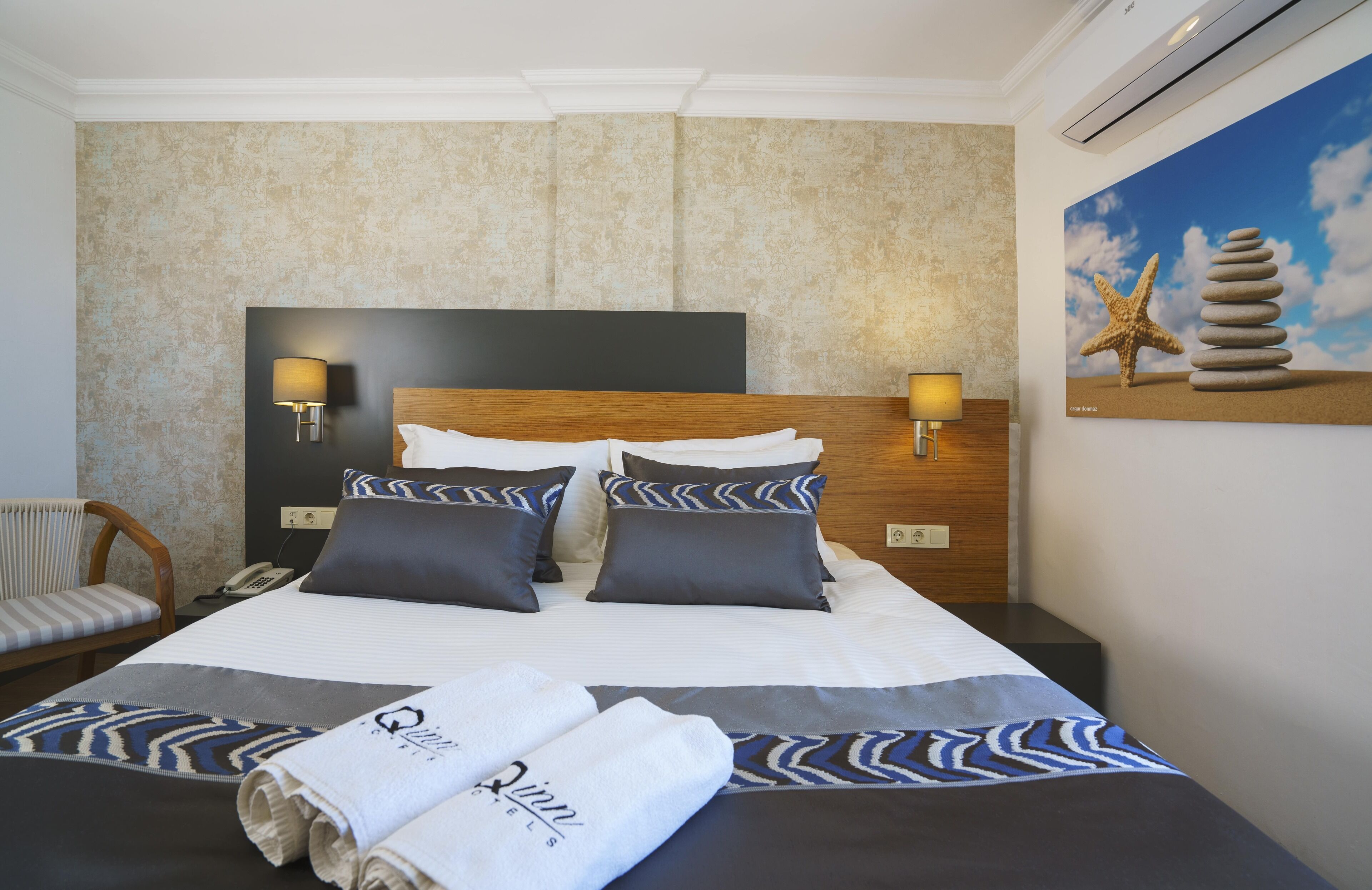 A hotel room with a large bed, two wall-mounted lamps, a wooden headboard, a painting of a starfish and stones, and a chair.
