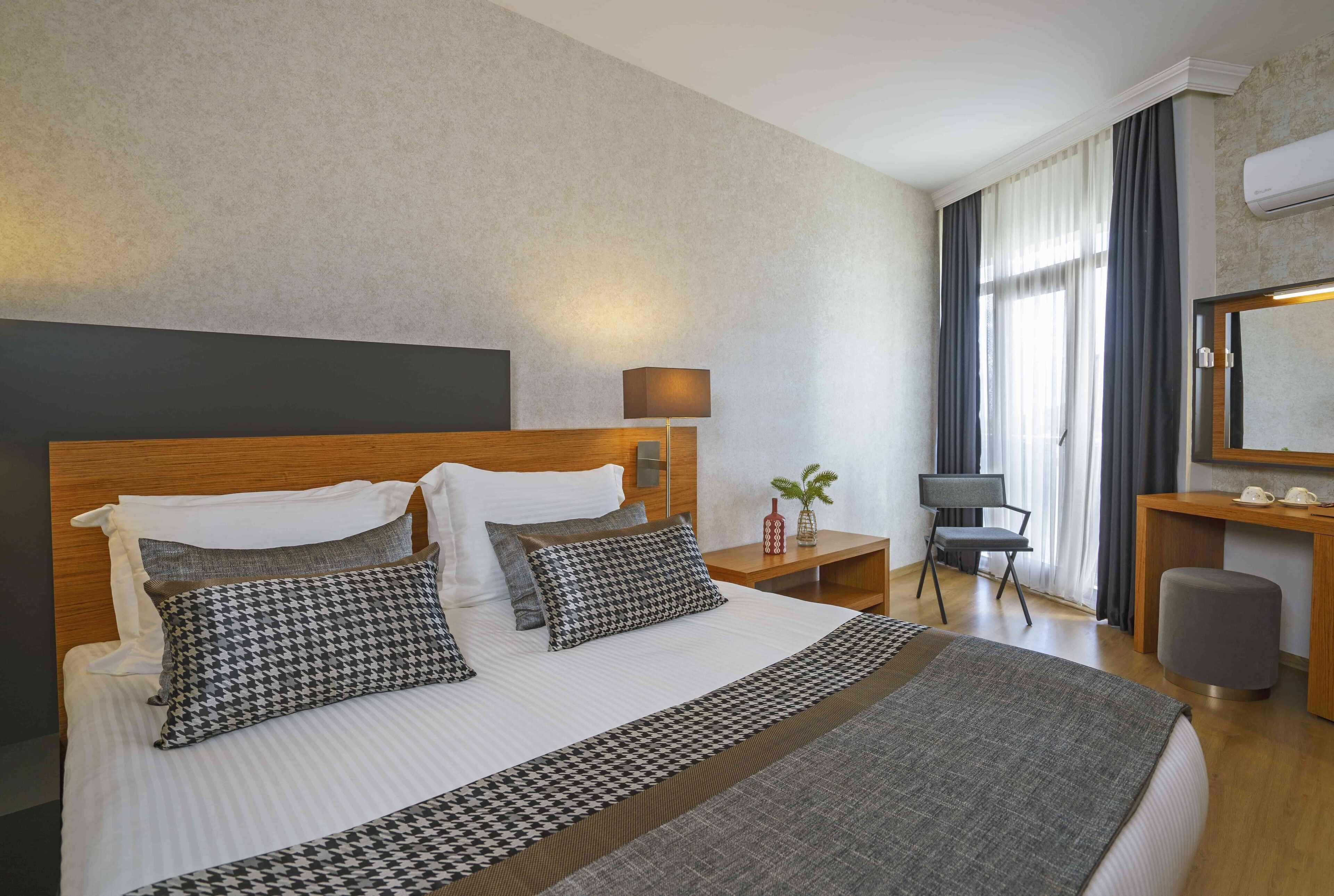 A hotel room with a large bed, a nightstand with a lamp, a chair, a desk with a mirror, and a window with curtains.