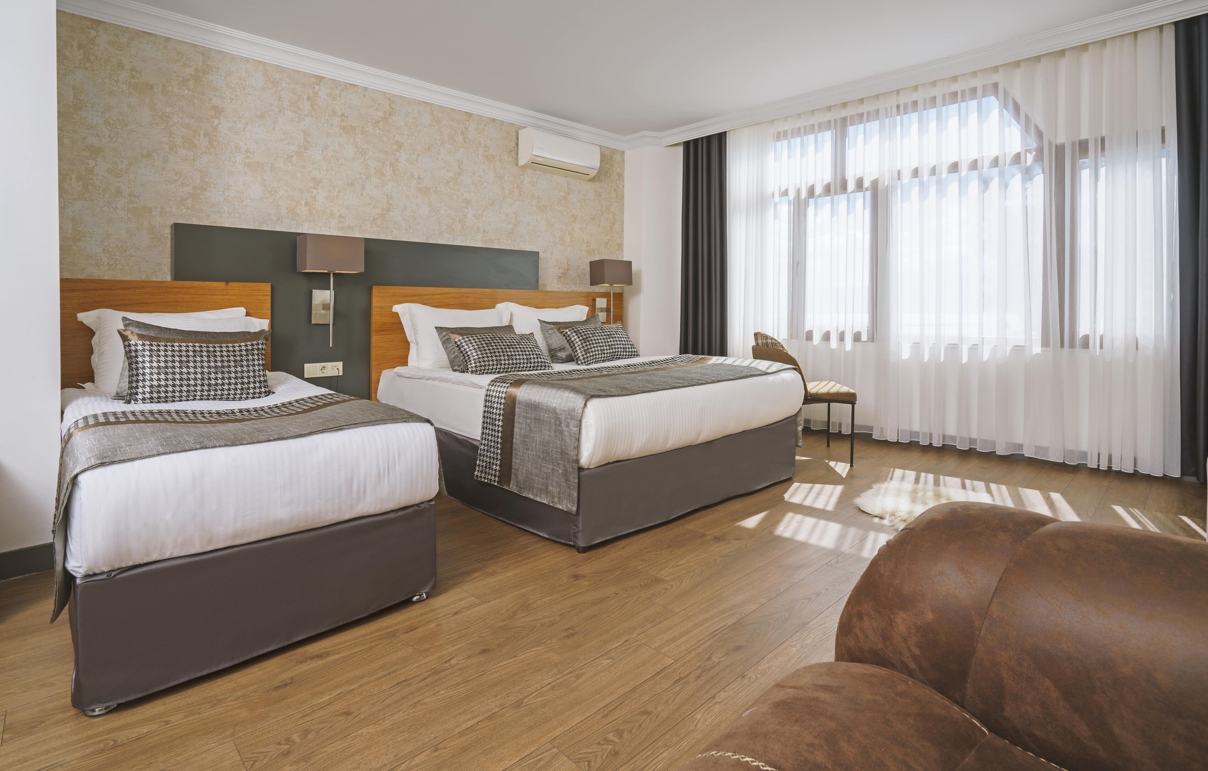 A hotel room with two beds, a wooden floor, a large window with curtains, and a chair.