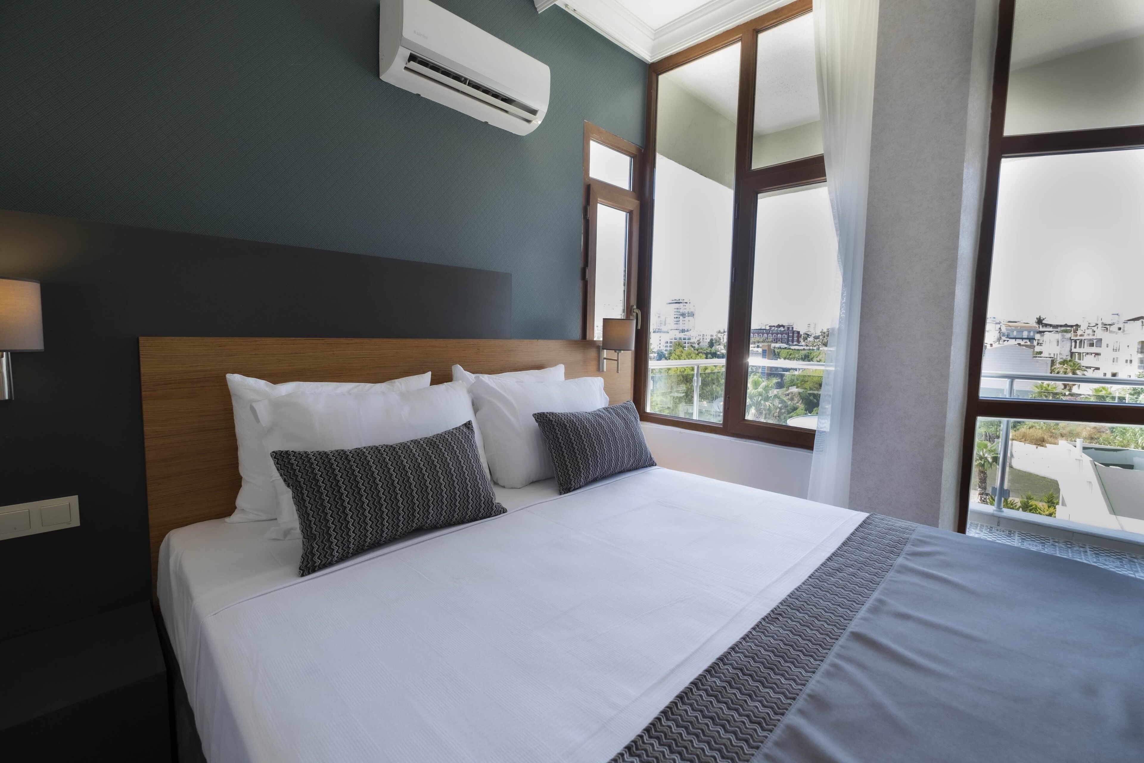 Hotellihuone, jossa on suuri sänky, ikkunasta avautuva näkymä kaupunkiin ja seinällä oleva ilmastointilaite.