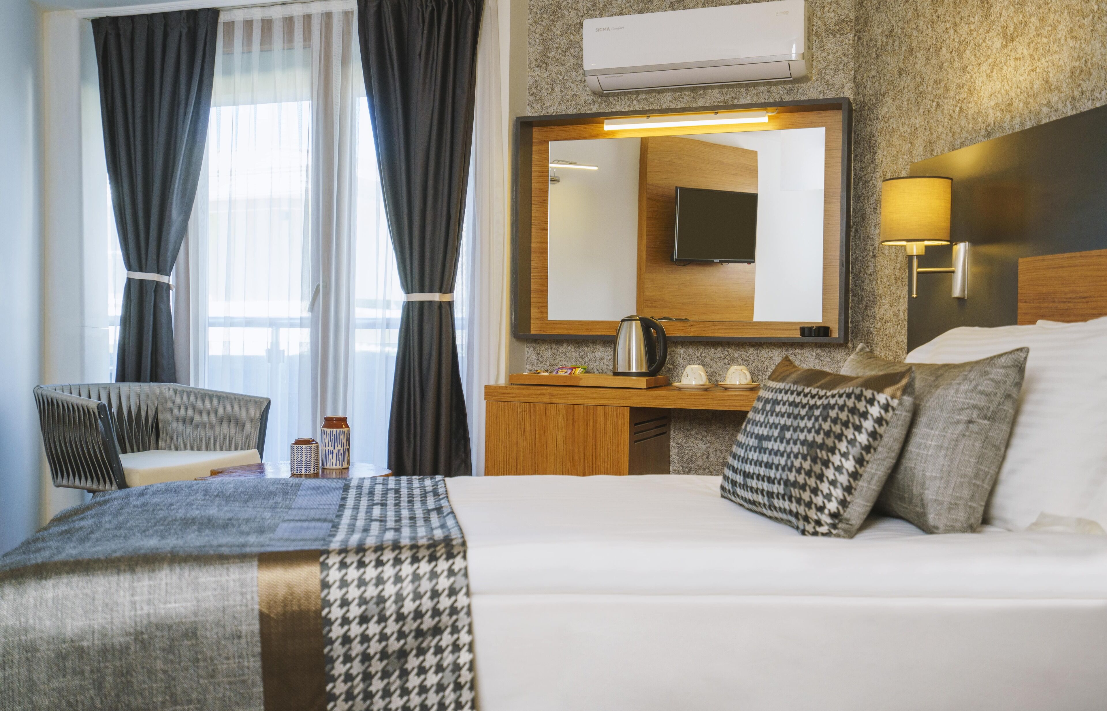 A hotel room with a large bed, a wooden desk, a mirror, a flat-screen TV, and a wall-mounted air conditioner.