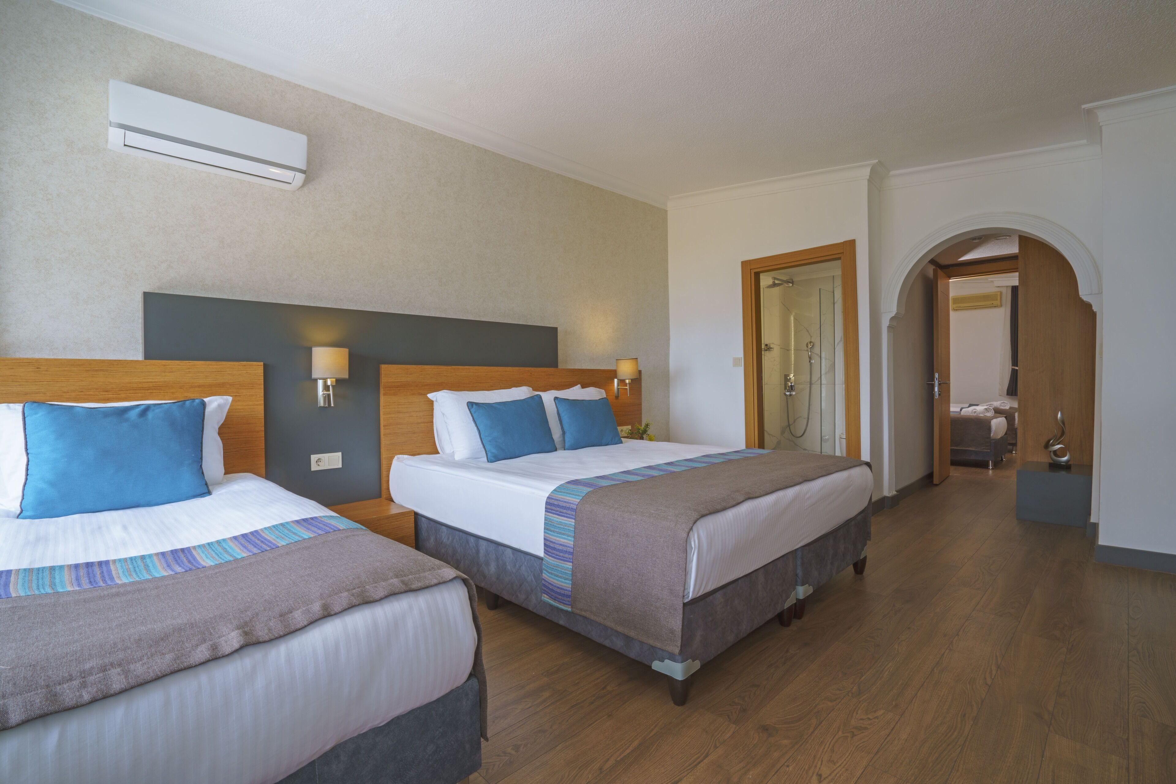 Une chambre d’hôtel avec deux lits, une tête de lit en bois, un climatiseur fixé au mur et un couloir menant à une autre chambre.