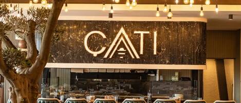 L’intérieur d’un restaurant au design moderne, avec des ampoules suspendues, un grand panneau lumineux portant l’inscription « CATI » et des sièges confortables recouverts d’un tissu vert moelleux.