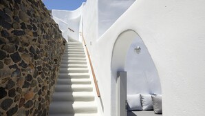 A narrow stairway flanked by white walls, a stone wall on one side, and a white door with a bench and pillows on the other. - El Verano Suites (Santorini)