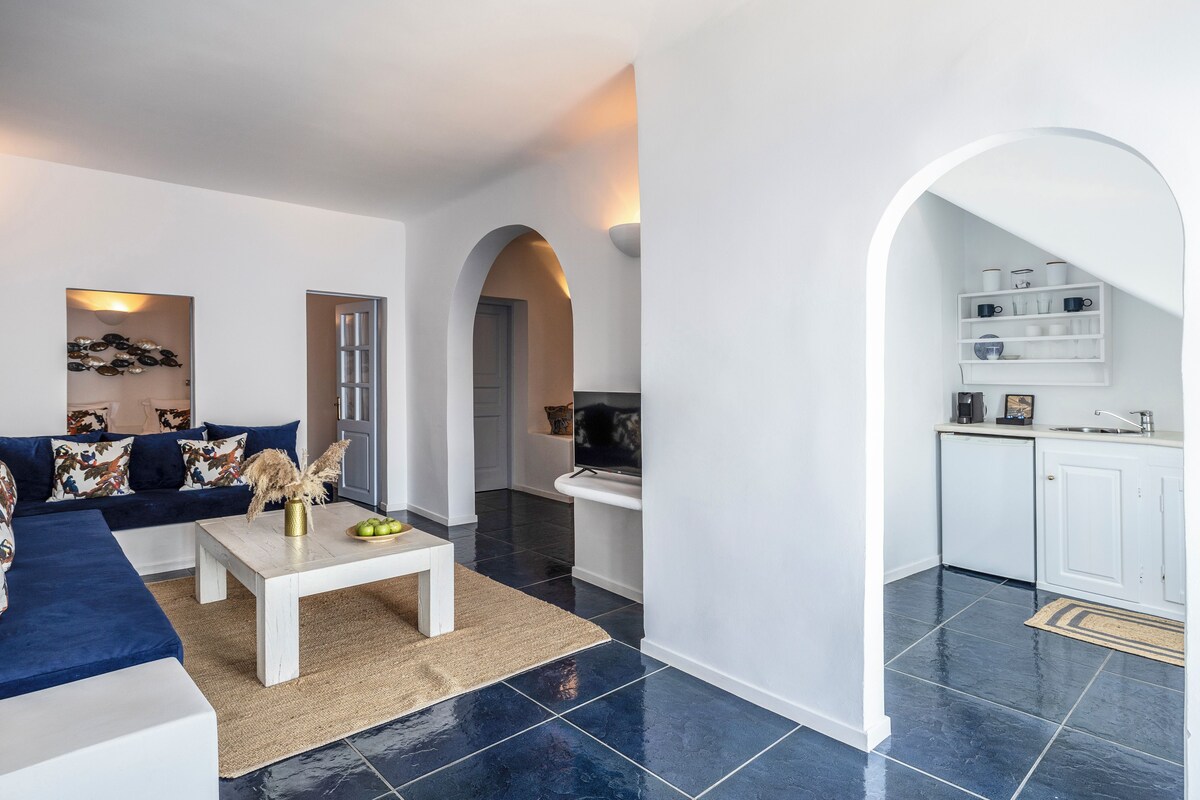 A modern living room with a blue sofa, a white coffee table, and a kitchen area with white cabinets and a sink.