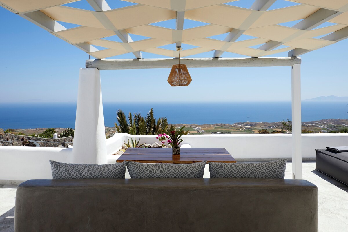 Un espacio de estar al aire libre techado con mesa y cojines, con vistas panorámicas al mar y al paisaje.