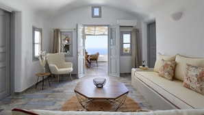 A spacious living room with a sofa, a chair, a coffee table, and a view of the sea through an open door. - El Verano Suites (Santorini)
