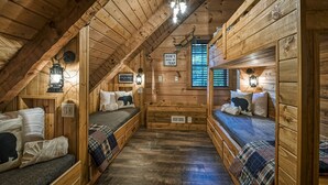 A cozy attic bedroom with wooden walls, a sloped ceiling, and a window. There are two beds with plaid bedding, a small table, and a sign that says "DON'T GROW UP E'T TRAP". - #1 KID'S CABIN!! JUST RENOVATED| CREEK| MINI GOLF⛳️PIRATE PLAY SHIP🏴‍☠️ HOT TUB (Cosby)