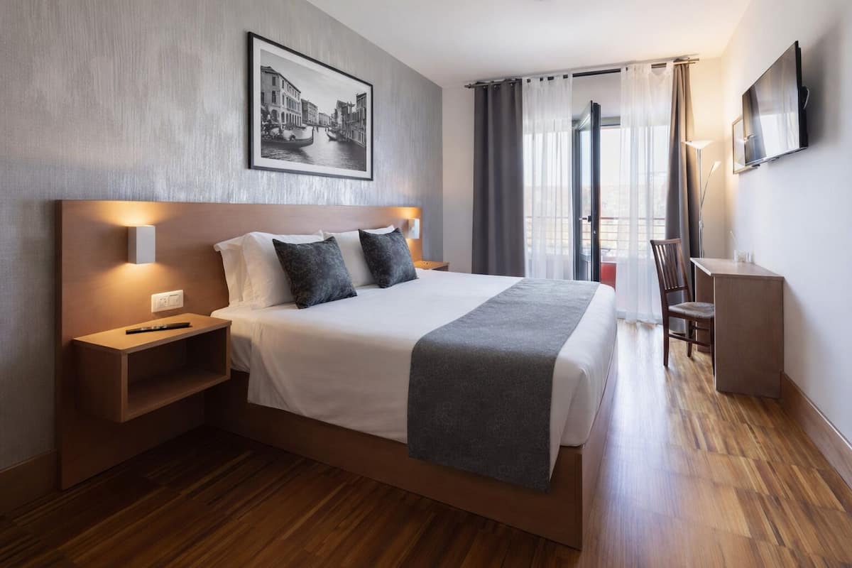 A modern hotel room with a large bed, bedside tables, a desk, a TV, and a window with curtains.
