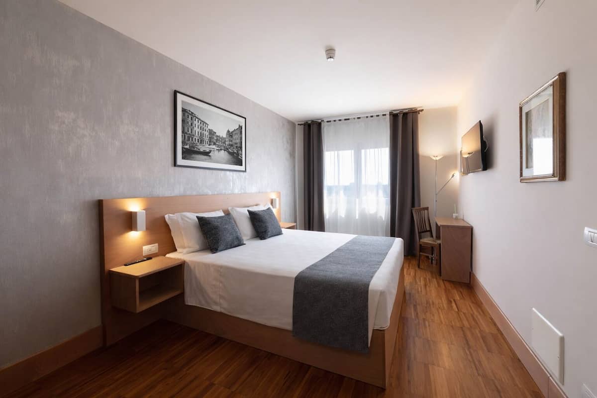 A modern hotel room with a large bed, bedside tables, a desk, a chair, a TV, and a framed picture on the wall.