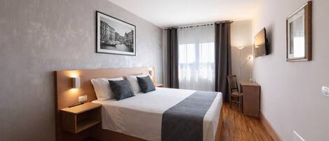 A modern hotel room with a large bed, bedside tables, a desk, a chair, a TV, and a framed picture on the wall.