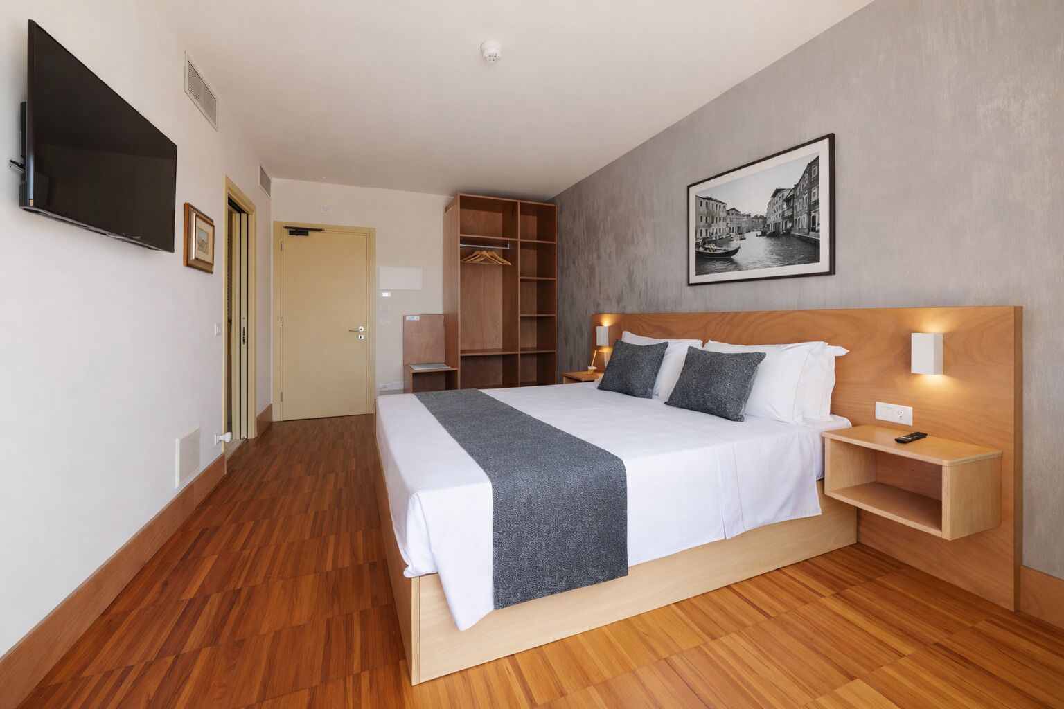 Una habitación de hotel moderna con una cama grande, suelo de madera, un armario, un televisor y un cuadro en la pared.